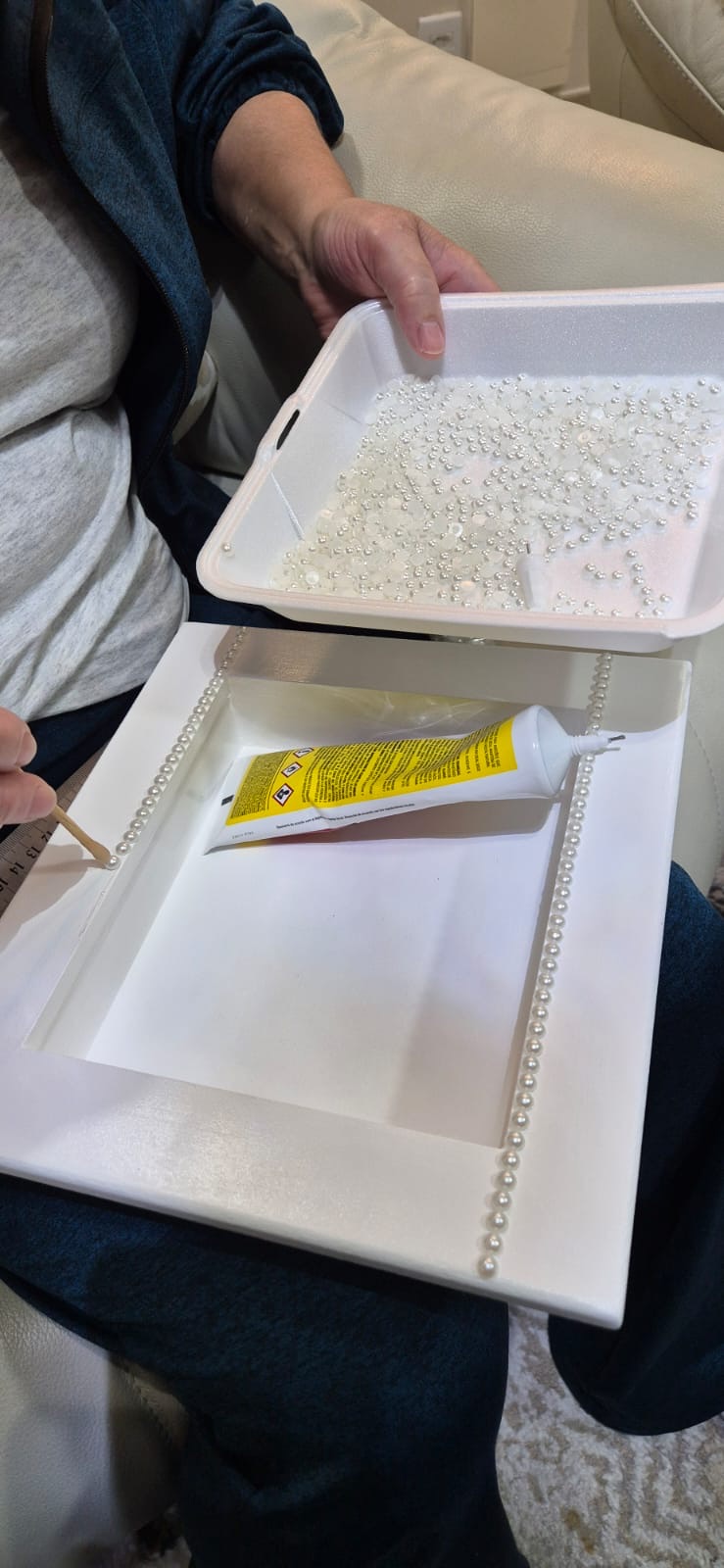 Mão da Cilene colando as pérolas uma a uma na borda do quadro, com o tubo de cola ao lado
